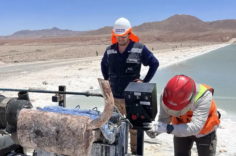 Dos operarios con casco instalan un panel de control y telemetría junto a una bomba diesel, a orillas de una poza del Salar de Atacama
